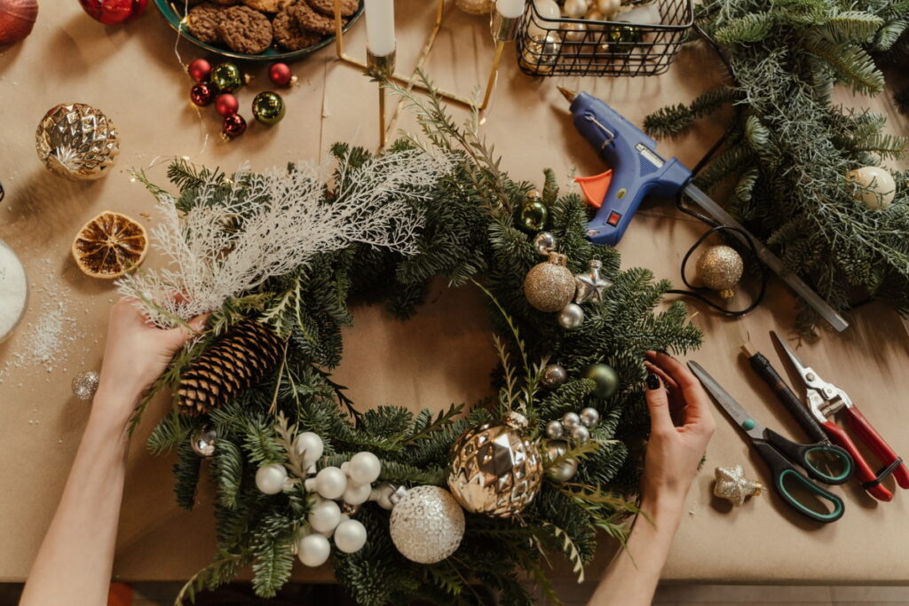 Hands decorating a handmade Christmas wreath with evergreen branches, pinecones and gold ornaments.