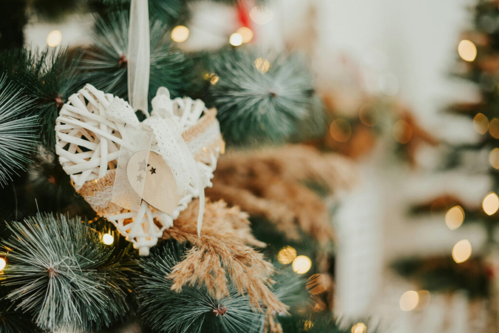 Handmade white woven heart ornament with lace ribbon hanging on a Christmas tree with natural decorations and warm lights.