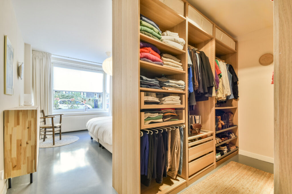 Organized wooden walk-in closet with folded clothes, hanging garments, and shoe shelves in a compact bedroom.