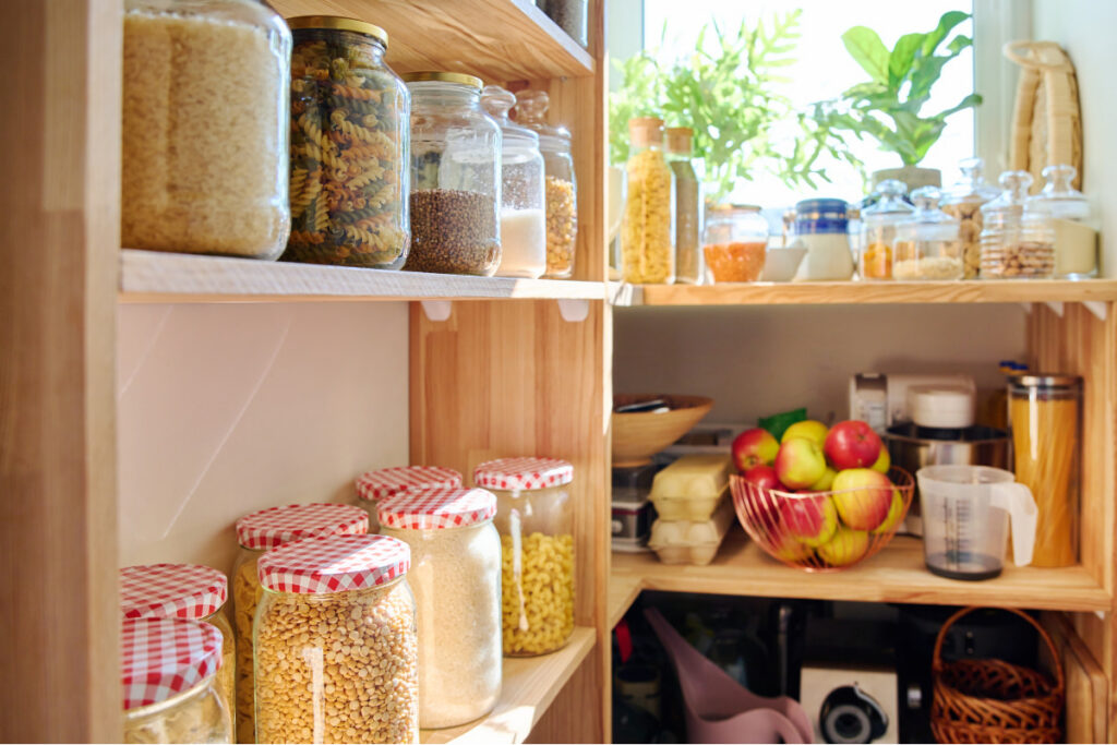 Open wooden pantry shelves with glass jars of dry foods, kitchen essentials, and fruit arranged for easy access.