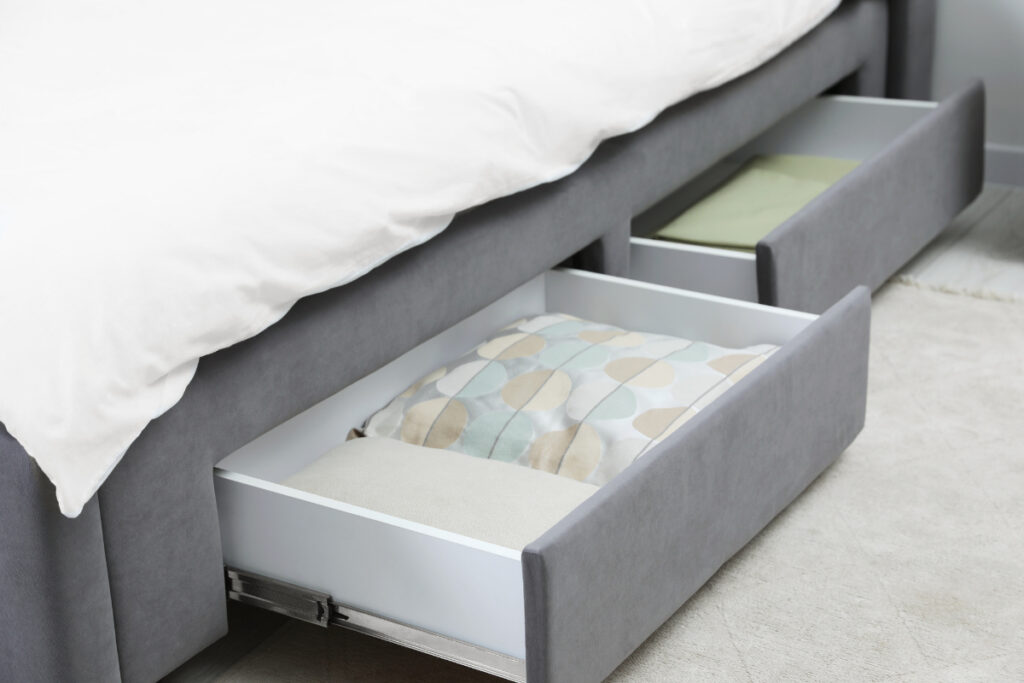 Gray upholstered bed with built-in storage drawers holding neatly folded bedding to maximize space in a small bedroom.